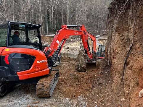 An orange excavator cutting into a steep earth bank beside a creek, with a second excavator working behind it.