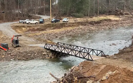 A black steel truss bridge spanning a wide creek in a wooded valley, with pickup trucks parked on the gravel road beyond it and storm debris piled along the near bank.