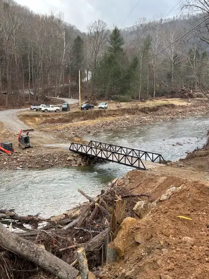 A black steel truss bridge spanning a wide creek in a wooded valley, with pickup trucks parked on the gravel road beyond it and storm debris piled along the near bank.