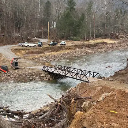 A black steel truss bridge spanning a wide creek in a wooded valley, with pickup trucks parked on the gravel road beyond it and storm debris piled along the near bank.