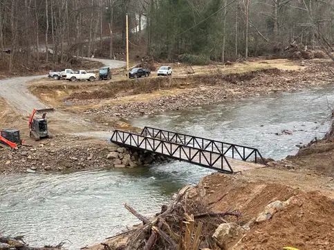 A black steel truss bridge spanning a wide creek in a wooded valley, with pickup trucks parked on the gravel road beyond it and storm debris piled along the near bank.