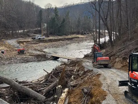 A flood-scoured creek valley with uprooted trees and broken timber piled along the bank, an excavator working the far side, and a damaged crossing in the middle distance.