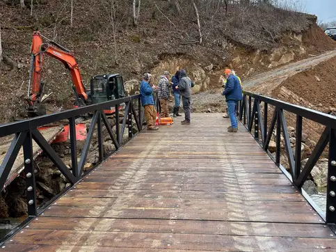 Looking along the timber deck of the installed bridge toward the far bank, where several people stand talking beside an excavator.