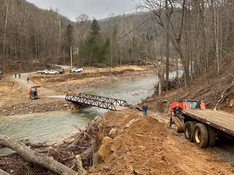 A flatbed trailer and a tracked loader on a cut earth approach above a creek, with the new bridge in place below and vehicles on the road beyond.
