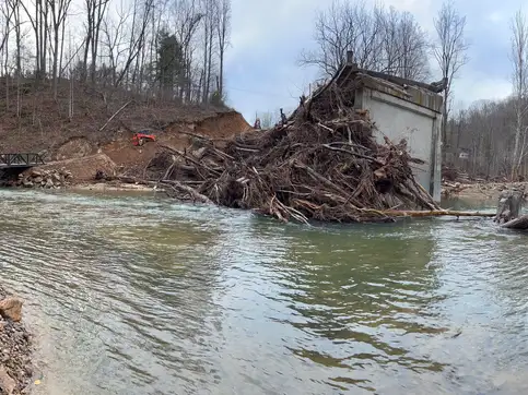 A wide view of a mountain river after the flood, with uprooted trees heaped against the stranded concrete abutment of the lost bridge and the new steel truss crossing in the distance at left.