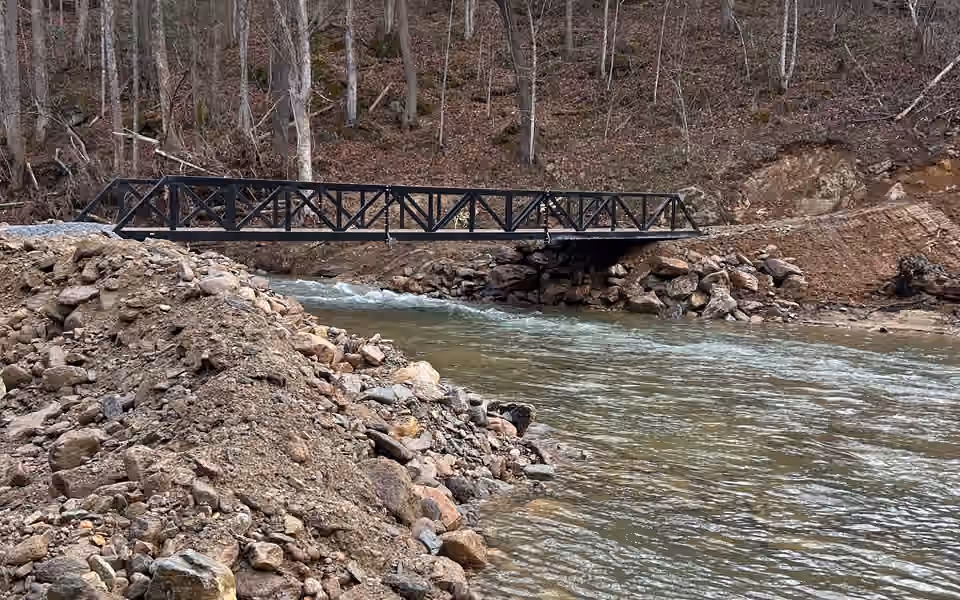A black steel truss bridge seen side on across a rocky creek, seated on stone abutments, in bare winter woodland.