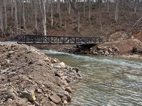 A black steel truss bridge seen side on across a rocky creek, seated on stone abutments, in bare winter woodland.