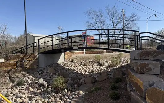 A black steel arched pedestrian bridge with a curved handrail, spanning a dry landscaped creek bed of river rock between stone-block abutments.