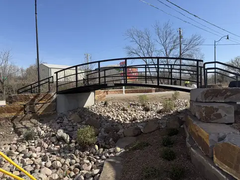 A black steel arched pedestrian bridge with a curved handrail, spanning a dry landscaped creek bed of river rock between stone-block abutments.