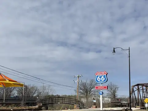 A wide view of the park with the black arched pedestrian bridge on the left, a Route 66 shield sign at the centre, and the weathered steel of the historic through-truss bridge on the right.