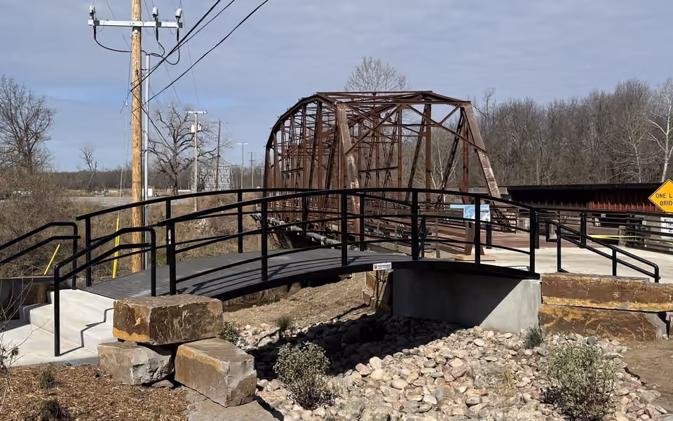 A new arched steel bridge with black railings on concrete abutments over a rock-lined swale, with the rusted truss of a historic highway bridge rising behind it.