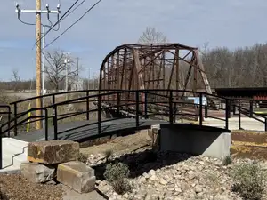 A new arched steel bridge with black railings on concrete abutments over a rock-lined swale, with the rusted truss of a historic highway bridge rising behind it.