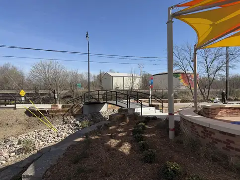 The arched pedestrian bridge seen across the park, with a brick plaza wall and an orange and yellow shade sail in the foreground.