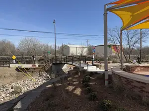 The arched pedestrian bridge seen across the park, with a brick plaza wall and an orange and yellow shade sail in the foreground.