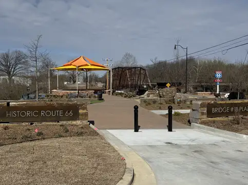 The park entrance, with carved stone signs reading Historic Route 66 and Bridge #18 Plaza either side of a brick walkway leading toward the historic truss.
