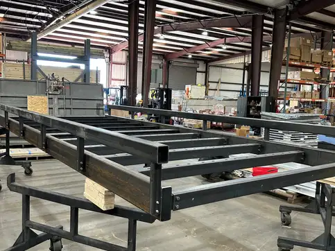 An uncoated steel bridge kit frame on shop trolleys, bare grey steel with a low bump rail down each side and no decking fitted.