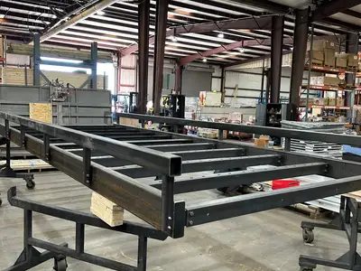 An uncoated steel bridge kit frame on shop trolleys, bare grey steel with a low bump rail down each side and no decking fitted.