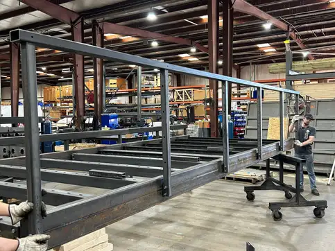 An uncoated steel bridge kit frame on shop trolleys with a full height handrail down one side, a worker steadying the far end.