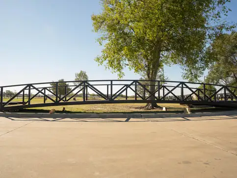 A long black arched steel truss span resting on timber blocks on the grass beside a concrete yard, under a spreading tree.