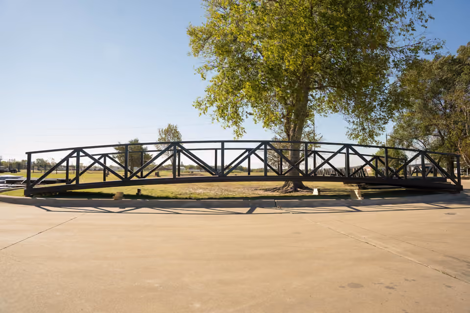 A long black arched steel truss span resting on timber blocks on the grass beside a concrete yard, under a spreading tree.