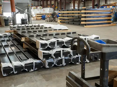 Bridge components wrapped and staged on a workshop floor, steel sections in plastic with stacked timber decking on racks behind.