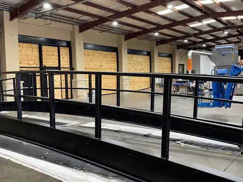 An arched bridge standing complete inside a workshop showroom, its deck and horizontal railing following the curve of the rolled beam beneath it.