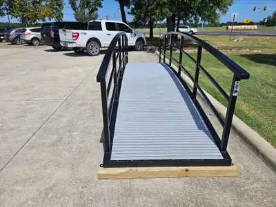 A short black arched steel bridge with tall curved handrails and a grey composite deck, set on timber on a concrete lot, with pickup trucks parked behind.