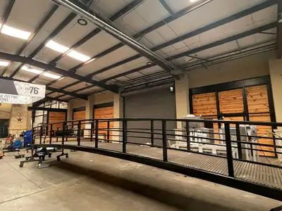 A long, low arched steel bridge with horizontal pipe rails and a dark composite deck, in a workshop.