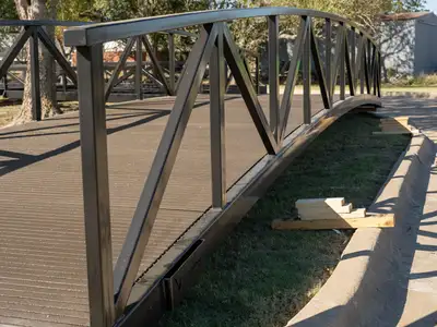 Looking along a black arched steel truss bridge with a dark composite deck, resting on the grass beside a curb, with trees behind.