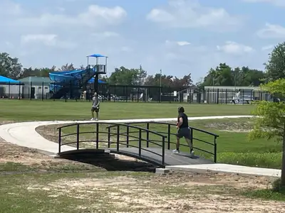 A black arched steel bridge with curved pipe handrails carrying a concrete path over a grassy swale in a park, with two people on the path and a play structure with a blue slide behind.
