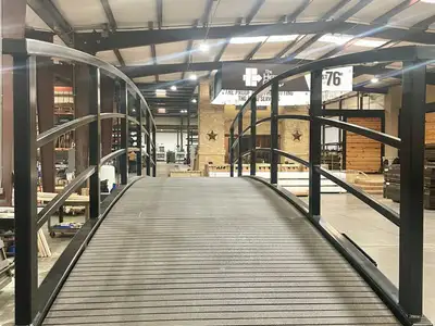 Looking along the dark composite deck of a black arched steel bridge with curved pipe handrails, standing on the floor of a workshop under bright ceiling lights.