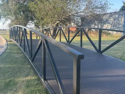 A long black arched steel truss bridge with a dark composite deck, on the grass beside a curving curb, with trees behind.