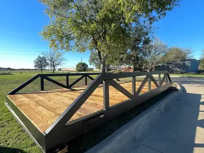 A short black steel truss bridge with a timber deck standing on timber blocks beside a curb, with a cast maker's plate on its side, under a tall tree and a clear sky.