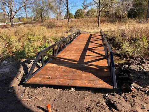A black steel bridge with low truss sides and a stained timber deck set across a dry creek bed, in brush under bare autumn trees.