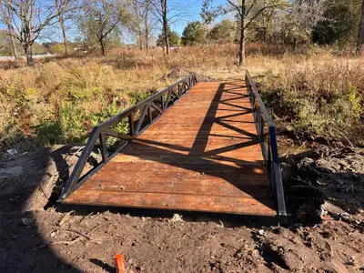 A black steel bridge with low truss sides and a stained timber deck set across a dry creek bed, in brush under bare autumn trees.