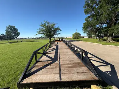 Looking down the length of a long black steel truss bridge with a timber deck, standing on blocks on the grass beside a drive, with trees and a clear sky beyond.