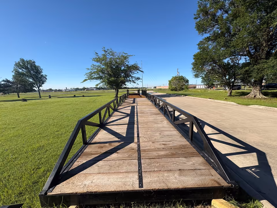 Looking down the length of a long black steel truss bridge with a timber deck, standing on blocks on the grass beside a drive, with trees and a clear sky beyond.