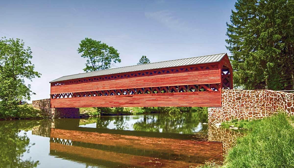 Covered pedestrian bridge made with wood trusses and a red finish over a creek.