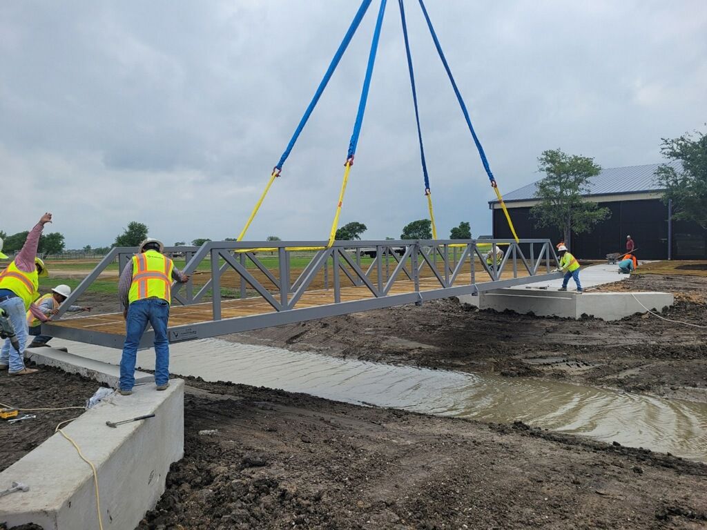 Crane lifting a fully assembled steel truss bridge into place over a creek, with installation crew positioning the span onto concrete abutments.