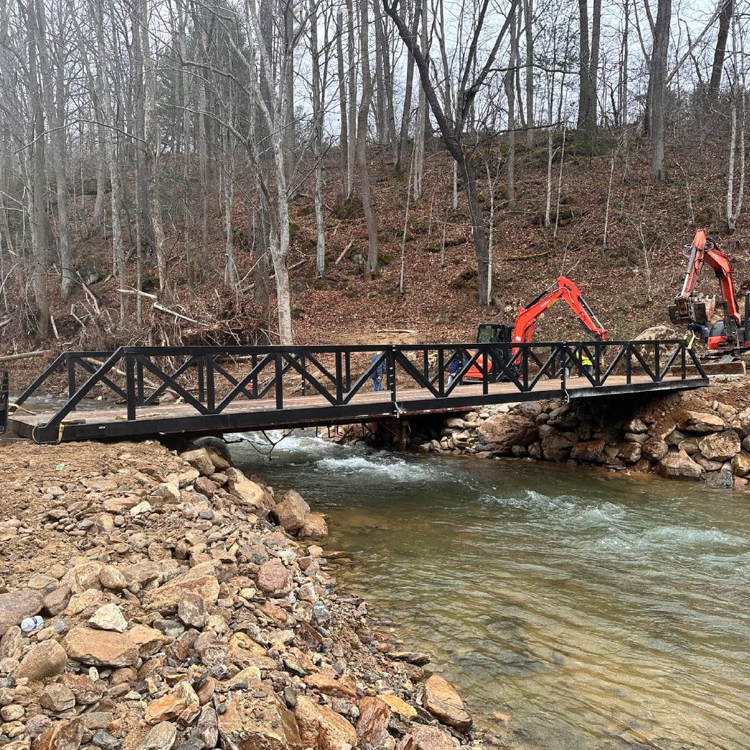 Roadrunner Bridge steel truss bridge being installed across a rocky creek using excavators and a skid steer in a wooded area.