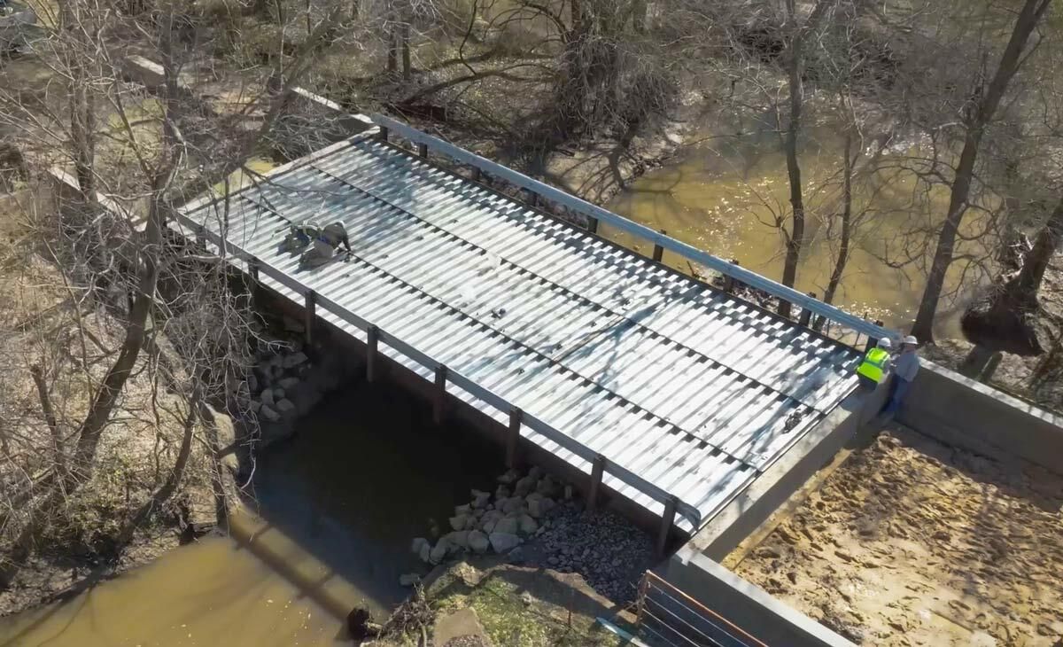 Galvanized modular truss pedestrian bridge over a creek with ongoing installation.