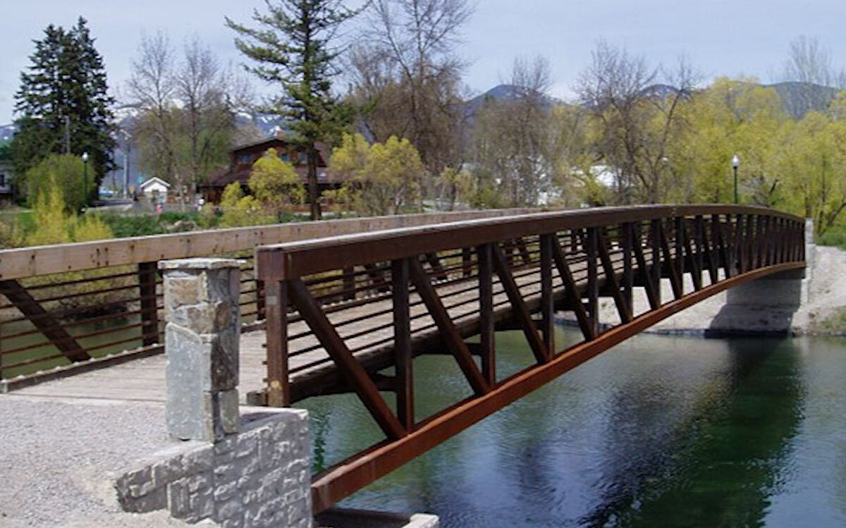 Warren truss pedestrian bridge with timber deck and steel railings over water.