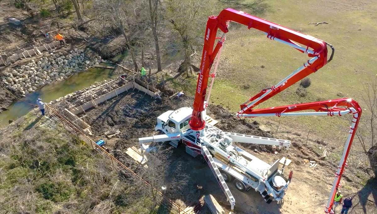 Warren truss bridge installation with concrete pump over creek site.