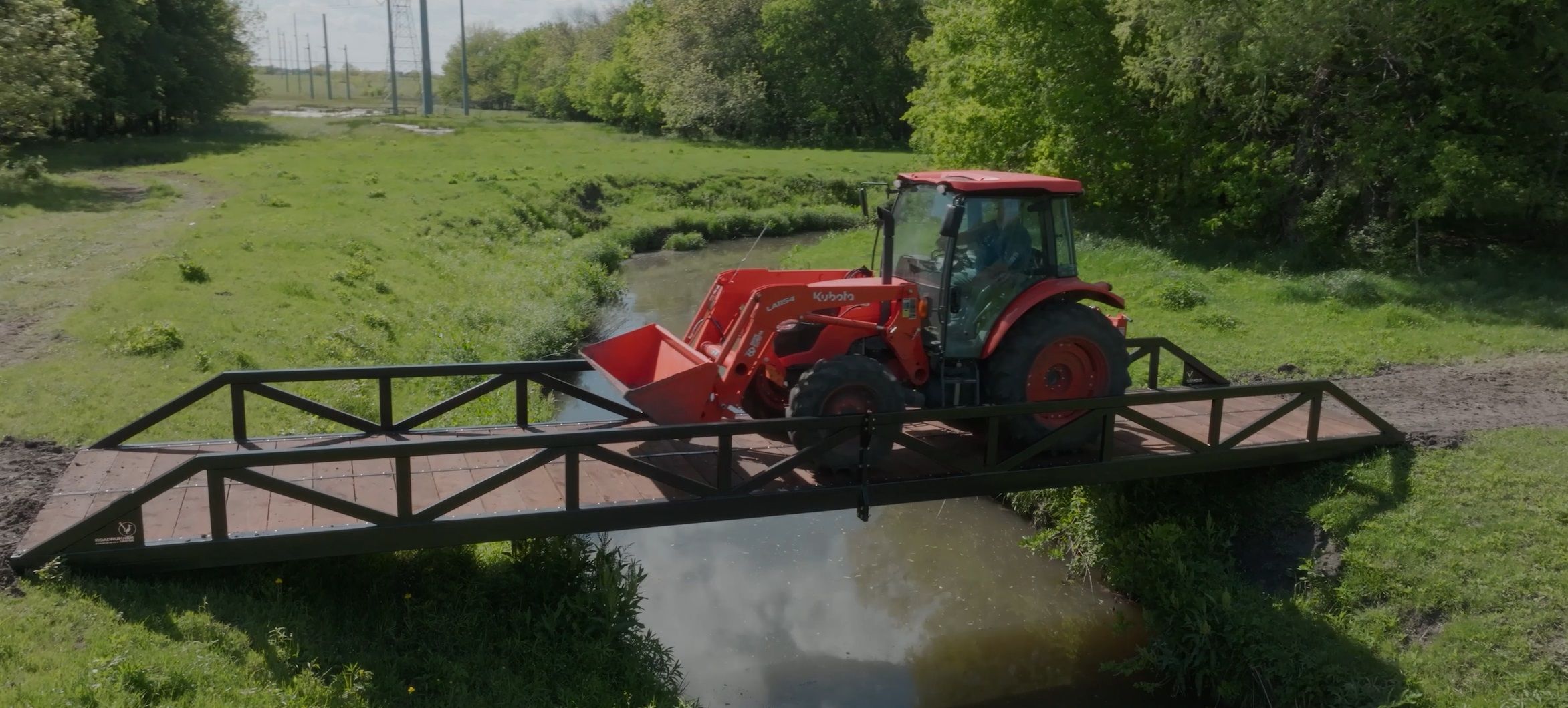 Pratt truss bridge supports tractor; painted with wooden deck; over creek.