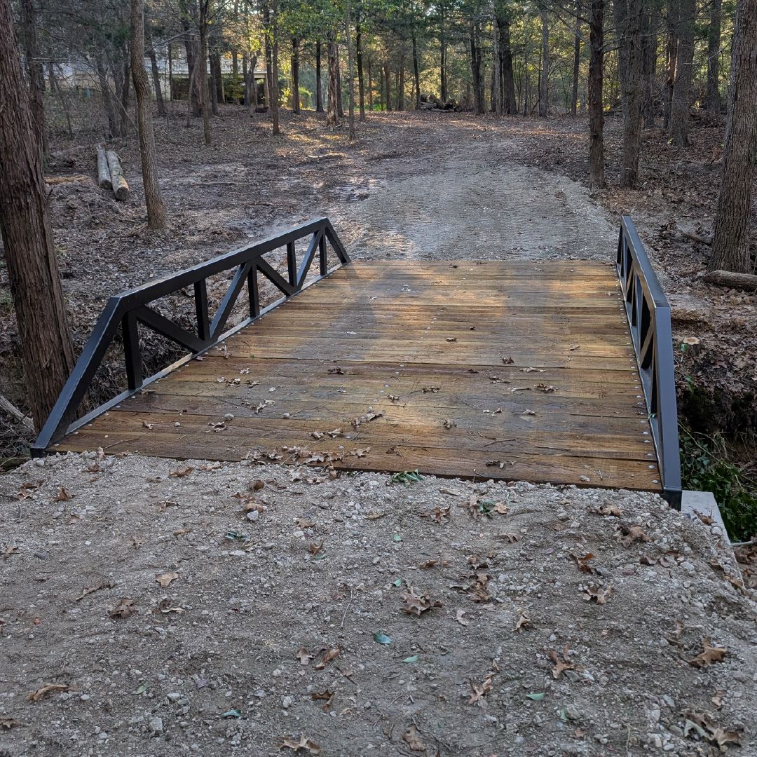 Roadrunner Bridge steel truss bridge with wood deck installed on a gravel driveway crossing a small creek in a wooded property.