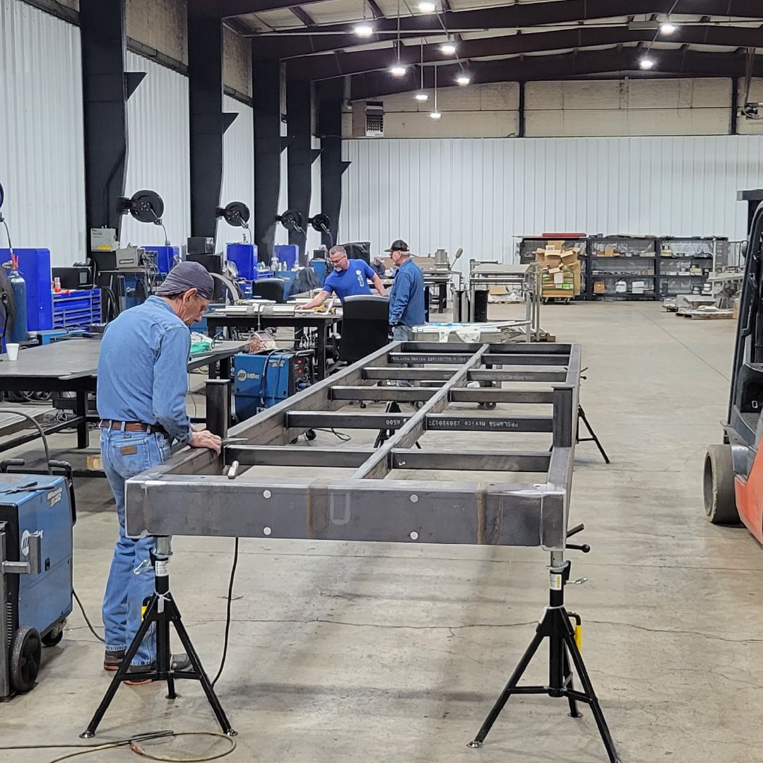 Steel bridge frame being welded and assembled by workers inside the Roadrunner Bridge fabrication shop.