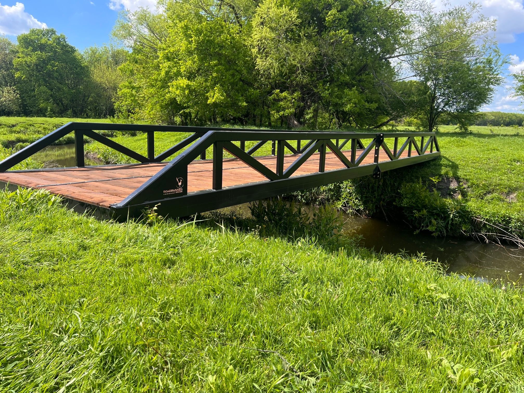 Pratt truss pedestrian bridge with painted finish and timber deck over a creek.