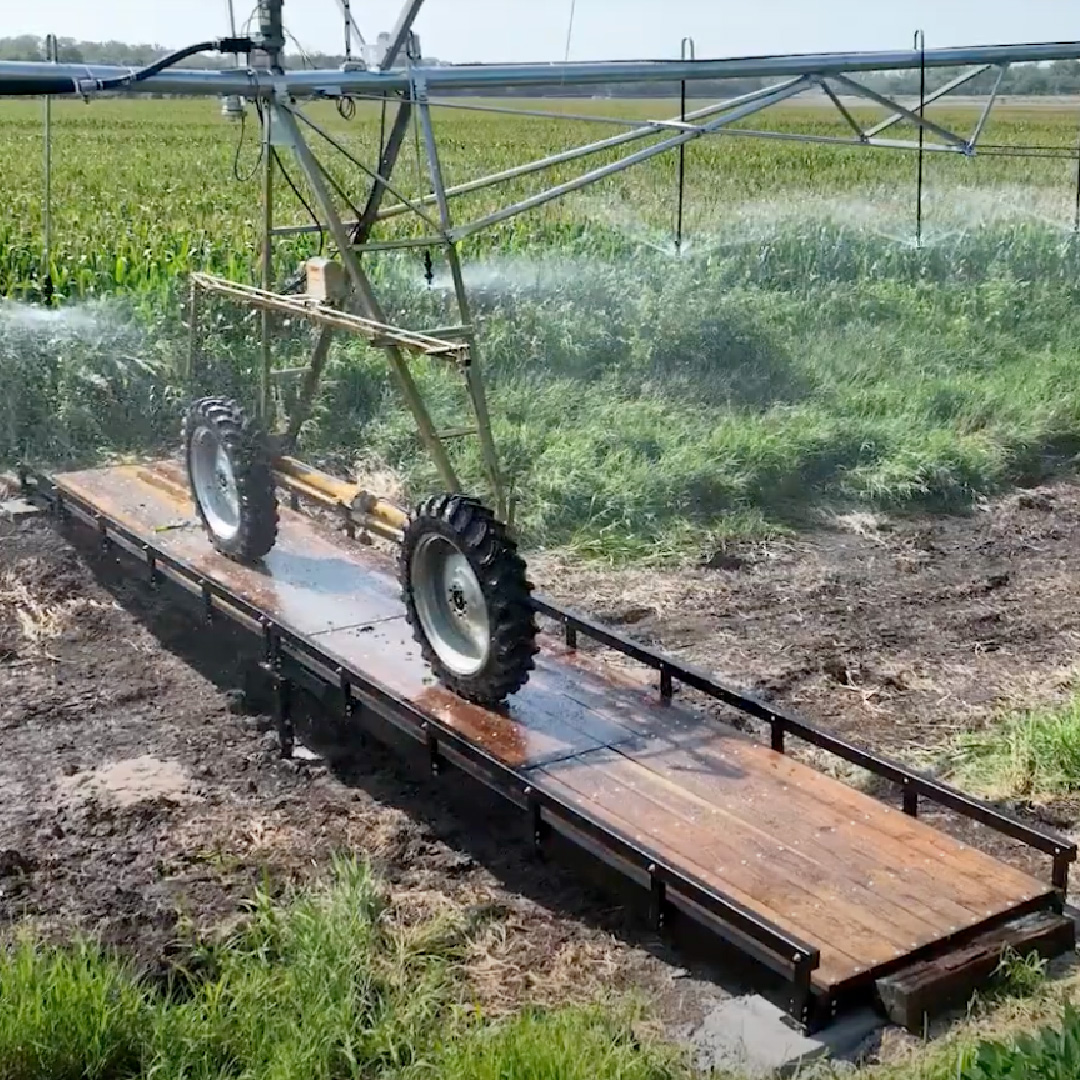 Center pivot irrigation system crossing a Roadrunner Bridge steel farm bridge over a muddy drainage ditch in an agricultural field.