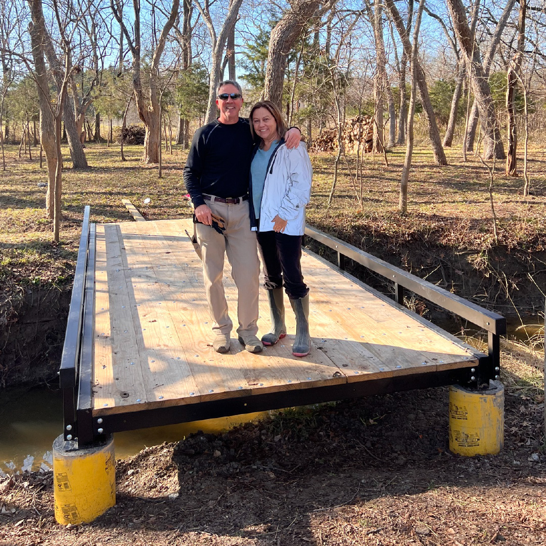 Couple standing on a newly installed Roadrunner Bridge steel pedestrian bridge spanning a small creek in a wooded property.